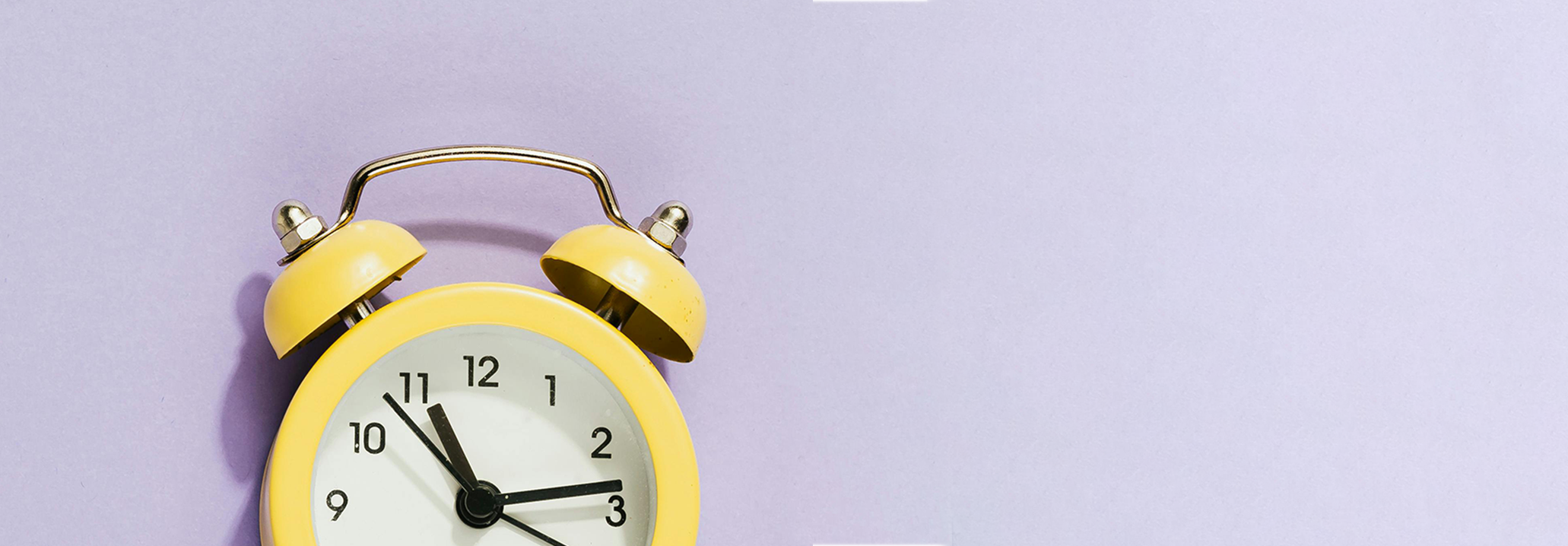 Close up of a yellow alarm clock against a plain lilac baclground