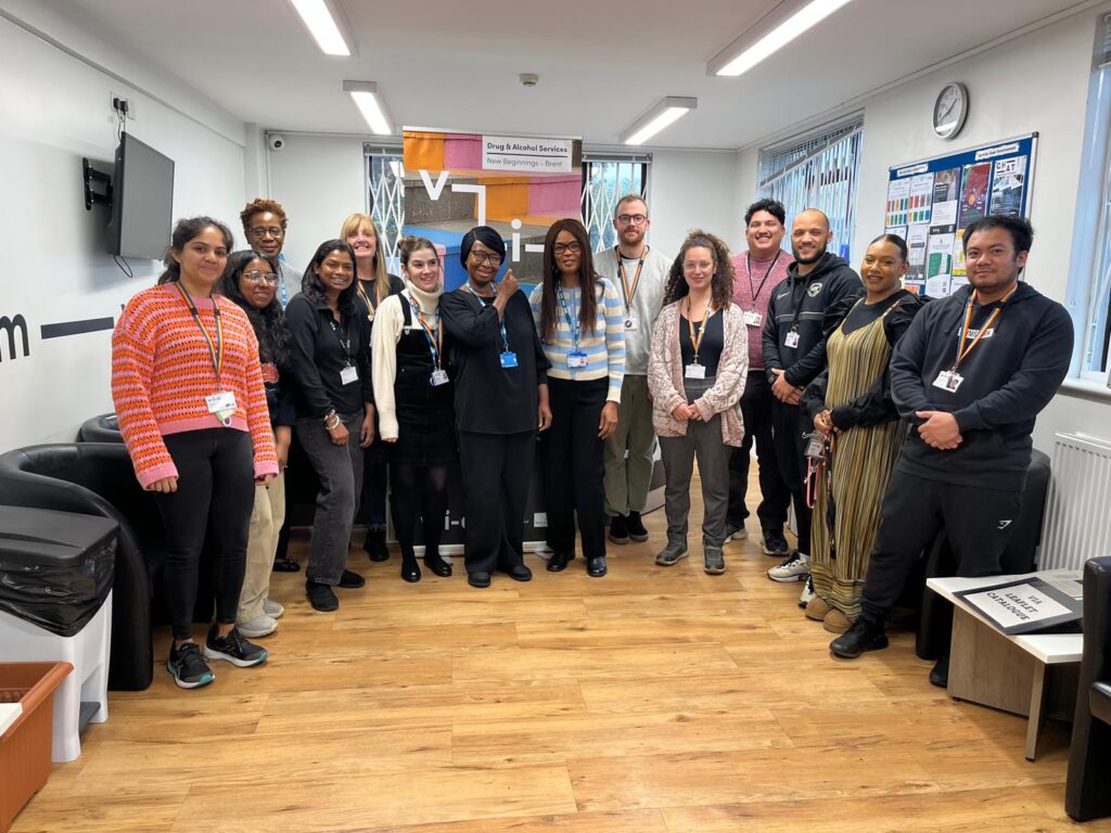 A group of 16 people standing in a room with wooden flooring and a welcome banner.