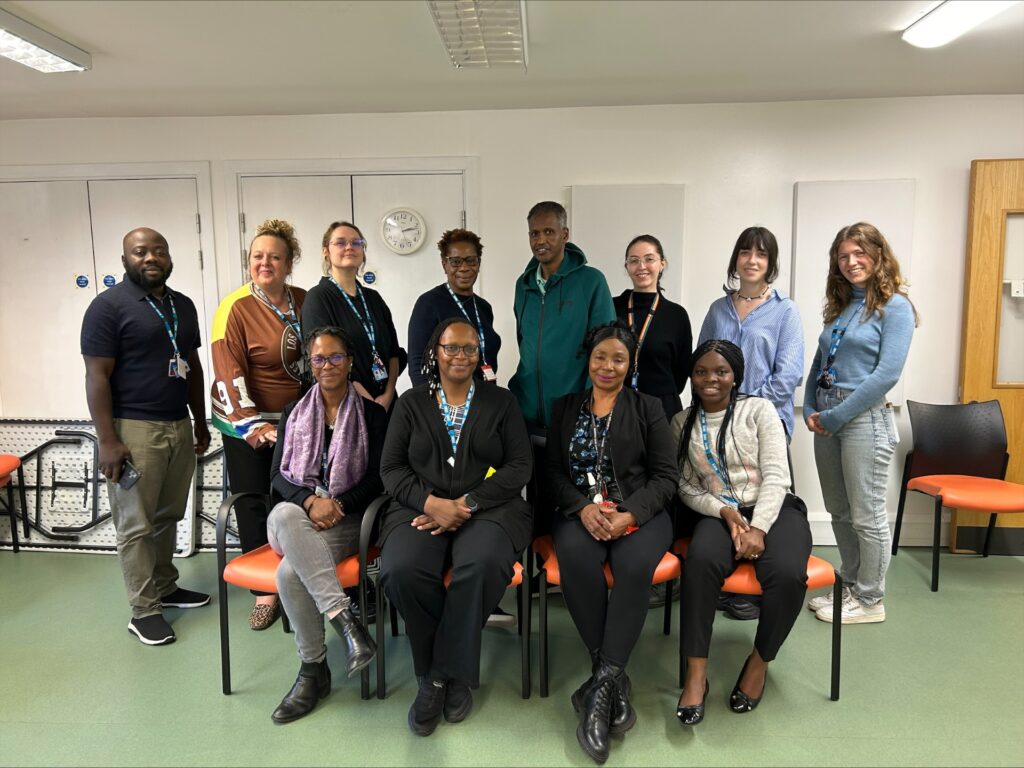 A group of 12 people gathered together in a room. They are posing for a photo, standing and sitting on orange chairs. 